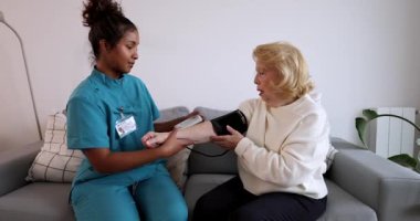 Ev ziyareti sırasında kanepede oturan yaşlı bir kadının kan basıncını ölçmek için sfenometre kullanan genç bir kadın bakıcı gerekli tıbbi yardım ve desteği sağlıyor.