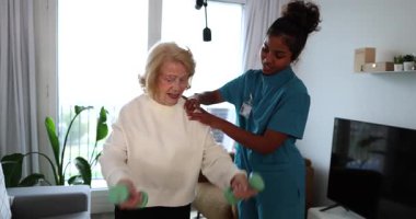 Fizyoterapi seansında kendini adamış bir kadın hemşirenin yardımıyla gülümseyen ve dambılları kaldıran yaşlı bir kadın aktif yaşlanma ve evde sağlıklı bir yaşam tarzını teşvik ediyor.