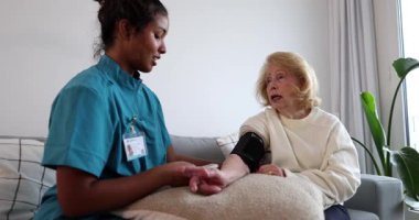 Genç bayan hemşire yaşlı bir kadına evde bakıcılık yapıyor. Konuşurken kanepede kan basıncını ölçüyor. Üst düzey sağlık hizmetleri ve tıbbi yardım kavramlarını temsil ediyor.