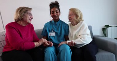 Dost canlısı çok ırklı kadın tıp uzmanları, yaşlı kadınlarla el ele tutuşuyor, huzur ve destek sağlıyor bir bakımevinde ya da ev ziyareti sırasında kanepede otururken.