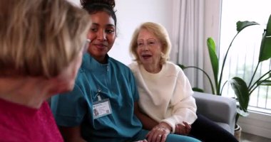 Genç bayan bakıcı, iki yaşlı hasta arasında bir kanepede oturuyor. Huzur, destek ve arkadaşlık sağlıyor. Huzurlu ve olumlu bir atmosfer yaratıyor.