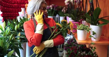 Çiçekçi dükkanından bir müşteriyle telefonda konuşurken elinde bir buket ayçiçeği tutan önlüklü ve eldivenli olgun bir çiçekçi.