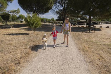 Anne ve kızı, yanlarında köpekleri ile güneşli bir çakıl yolunda el ele tutuşmuş yeşil ağaçların ve kuru çimenlerin arasında tasasız bir yaz tatili geçiriyorlar.