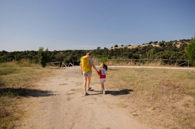 Kadın ve küçük kız el ele tutuşuyorlar, sırt çantaları takıyorlar ve yürüyüş kıyafetleri giyiyorlar, açık mavi gökyüzünün altında tozlu doğal bir patikada yürüyorlar, aile bağlarını ve ortak maceralarını gösteriyorlar.