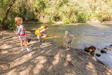 Aralarında bir yetişkin, çocuk ve evcil köpekleri de var. Nehir kıyısında su sıçratma ile etkileşim halindeler. Birlikte doğa aktivitelerinin tadını çıkarıyorlar.