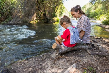 Anne ve kızı yemyeşil bir doğal ortamda akan bir nehrin kıyısında birbirlerine bağlanıyorlar. Çocuk çizim kitabına odaklanırken, ebeveyn de açık havada huzurlu bir anı paylaşıyor.