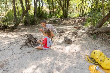 Genç anne kızına kamp ateşi için çıra ve dallardan çadır yapmayı öğretiyor. Açık havada hayatta kalma becerilerini ve güneşli ormanda neşeli aile bağlarını paylaşıyor.