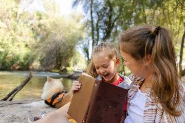 Mutlu anne ve kız nehir kıyısında dinleniyor, ilginç bir kitap okurken birlikte güzel vakit geçiriyorlar, ve köpekleri arkadan izliyor.