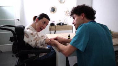 Genç erkek bakıcı, beyin felci geçirmiş bir kadına ev yardımı ve fiziksel terapi sağlıyor. Parlak, modern bir oturma odasındaki masada kol hareketleri ve egzersizlerine yardım ediyor.