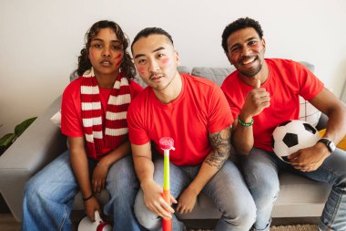 Kırmızı tişört ve yüz boyası giyen bir grup arkadaş, spor aksesuarlarıyla kutlama yaparken evdeki kanepede heyecanlı bir futbol maçı izliyorlar.