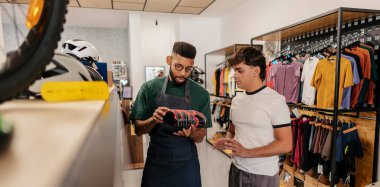 Bir erkek müşteriye bisiklet ayakkabısı özelliklerini açıklayan satış asistanı, aktif giyim ve ekipman dolu modern bir spor mağazasında uzman tavsiyesi sunuyor.