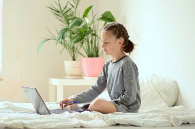 Genç bir liseli kız evdeki yatakta oturarak ev ödevini yapıyor. Kızın önünde bir laptop var..