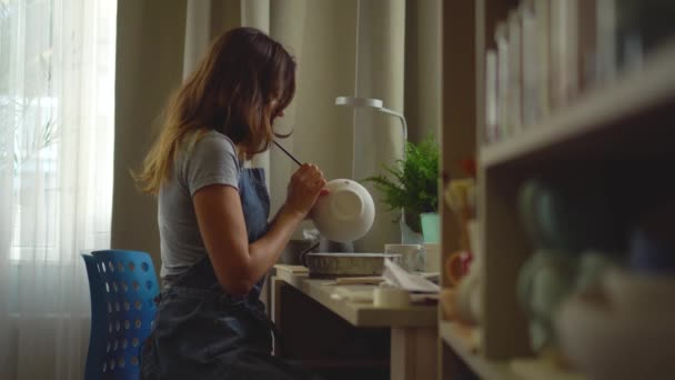 Vrouw die keramiek en aardewerk servies maakt in de werkplaats. jong meisje verft aardewerk met een borstel — Stockvideo