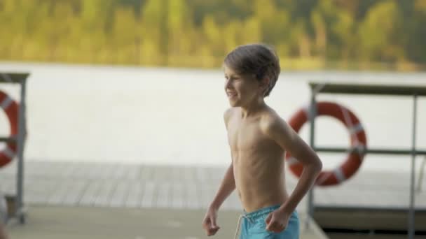 Dziewczyna i mały chłopiec w kombinezonie skaczący do jeziora z drewnianego molo. Zabawa w letni dzień. — Wideo stockowe