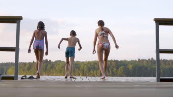 La familia feliz tiene vacaciones en el lago. Madre con hija e hijo saltan juntos desde el muelle. — Vídeos de Stock