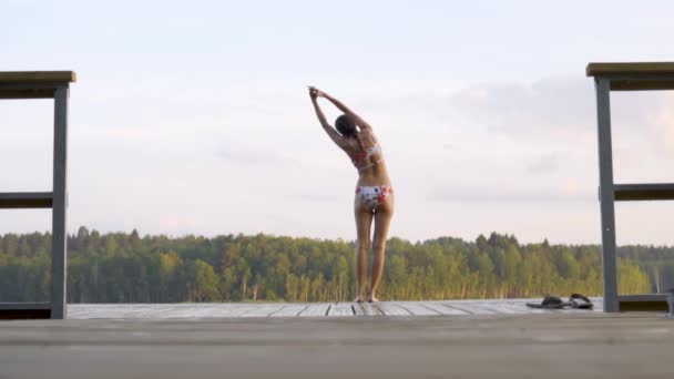 Una hermosa mujer se estira en el muelle. La chica se estira antes de nadar en el lago. — Vídeos de Stock