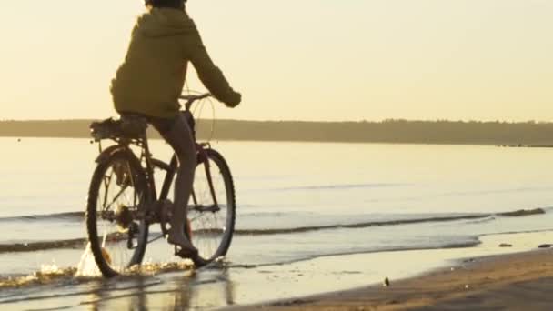 Dziecko jeździ na rowerze po wodzie w promieniach zachodzącego słońca wzdłuż plaży. Zajęcia sportowe o zachodzie słońca. — Wideo stockowe