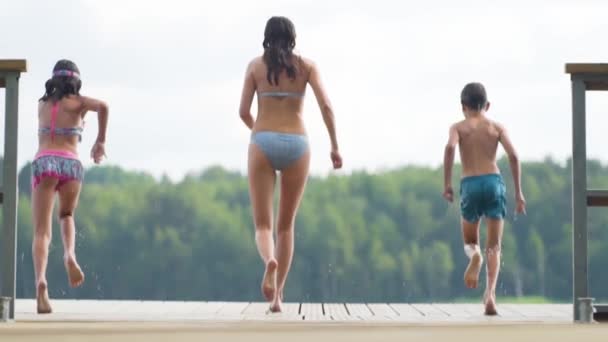 La familia feliz tiene vacaciones en el lago. Madre con hija e hijo saltan juntos desde el muelle. — Vídeo de stock