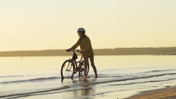 Il bambino va in bicicletta sull'acqua sotto i raggi del sole al tramonto lungo la spiaggia. Attività sportive al tramonto. — Video Stock