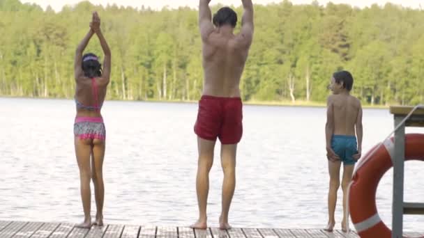La familia feliz tiene vacaciones en el lago. Padre con hija e hijo saltan juntos desde el muelle. — Vídeos de Stock
