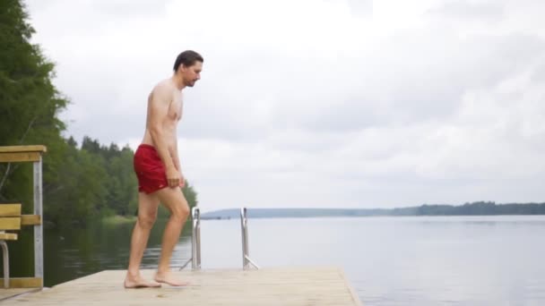 Giovanotto che salta nel lago dal molo di legno. Divertirsi nella giornata estiva. — Video Stock