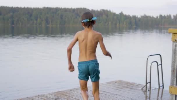 Mały chłopiec w kombinezonie skaczący do jeziora z drewnianego molo. Zabawa w letni dzień. — Wideo stockowe