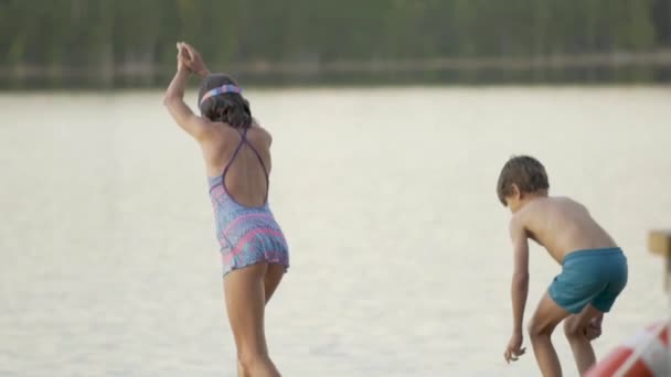 Niña y niño en traje de neopreno saltando al lago desde el muelle de madera. Divertirse en el día de verano. — Vídeos de Stock