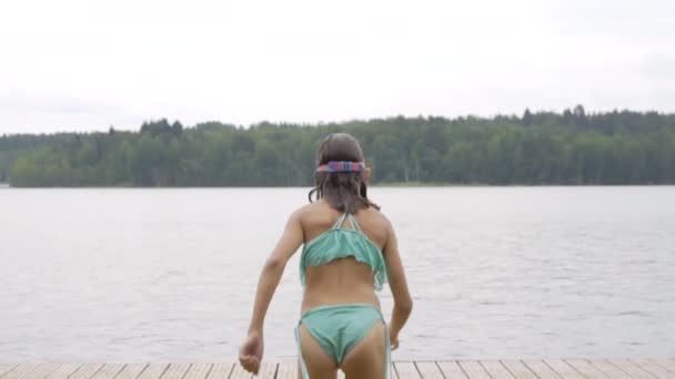 Dziewczyna w kombinezonie skacząca do jeziora z drewnianego molo. Zabawa w letni dzień. — Wideo stockowe