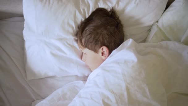 Søvnig dreng vågner tidligt om morgenen, strækker sig og gaber i sengen efter en sød drøm. Barn under dæksler, kigger på kameraet – Stock-video