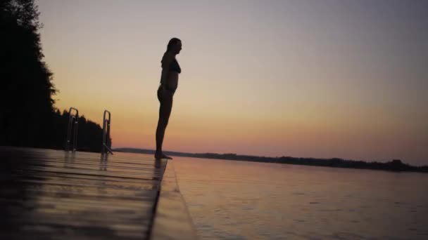 Een vrouw die van een houten pier in het meer springt. Plezier hebben op zomerdag. Jong meisje duikt in het water vanaf de steiger. — Stockvideo