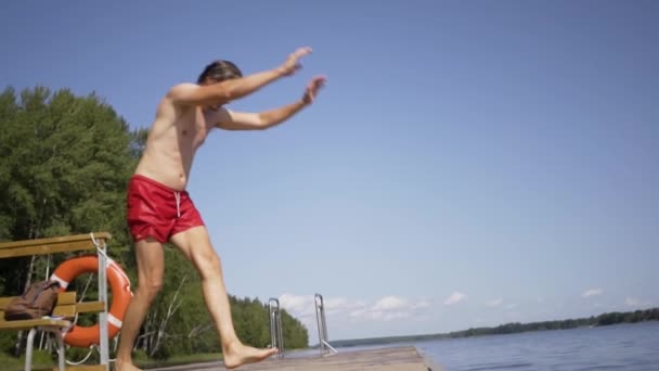 Mladý muž skáče do jezera z dřevěného mola. Bavíte se v letní den. — Stock video