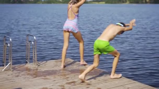 Mädchen und kleiner Junge im Neoprenanzug springen von einem Holzsteg in den See. Spaß am Sommertag. — Stockvideo