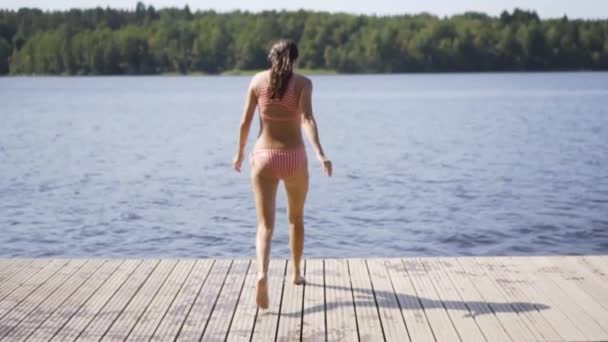 Mujer saltando al lago desde el muelle de madera. Divertirse en el día de verano. Chica joven sumergiéndose en el agua desde el muelle. — Vídeo de stock