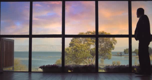 身穿西服的成年人在日落时走在一个大窗前 等待着某个人 凝视着湖面 动作慢 镜头长 — 图库视频影像