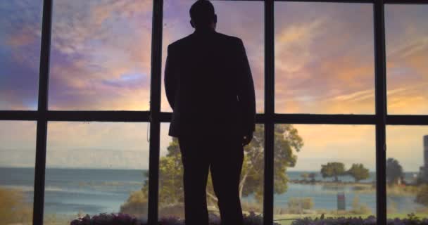 スーツの男は 湖を見て 夕日で大きな窓に向かって歩いて 彼の携帯電話を確認します スローモーション ロングショット — ストック動画