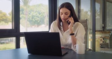 Ofiste büyük pencereli telefonla konuşurken içki içen bir iş kadını. Ortalama çekim, yavaş çekim.