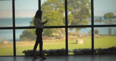 İş kadını, göl manzaralı büyük bir pencerenin yanındaki telefonunu kontrol ediyor. Yavaş çekim, uzun atış..