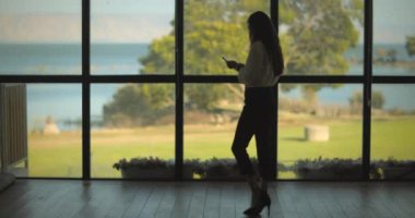 İş kadını, göl manzaralı büyük bir pencerenin yanındaki telefonunu kontrol ediyor. Yavaş çekim, uzun atış..