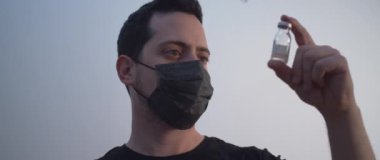 Siyah maskeli bir adam bir şişe aşıyı kaldırıyor ve ona bakıyor.