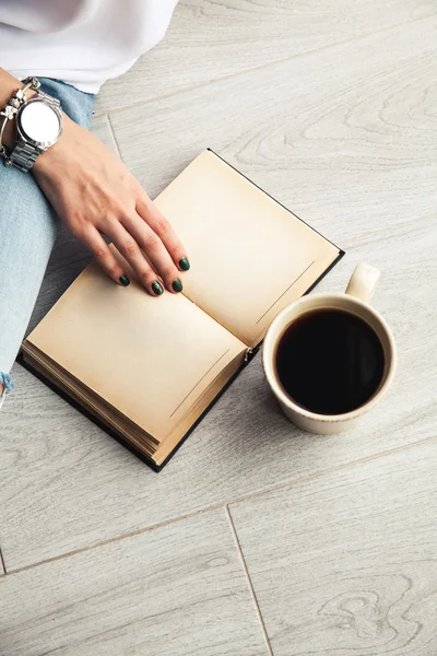 Jovem menina moderna em jeans rasgado lendo um livro com uma grande xícara de — Fotografia de Stock