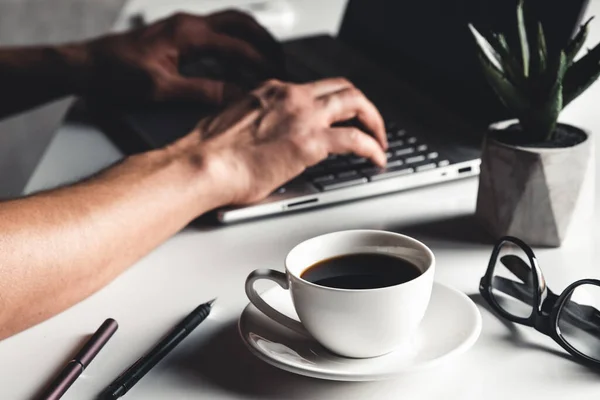 Un uomo scrive su un computer portatile, concetto di business, occhiali, una tazza di caffè e una penna su uno sfondo grigio. — Foto Stock