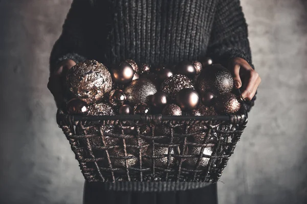塞满了圣诞或新年金银财宝装饰玩具的铁丝篮子，手握灰色保暖毛衣的女士手中 图库照片