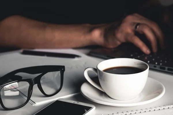 Un uomo scrive su un computer portatile, concetto di business, occhiali, una tazza di caffè e una penna su uno sfondo grigio. — Foto Stock