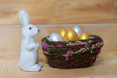 Tahta bir arka planda, yumurta sepetinin yanında beyaz bir oyuncak tavşan.