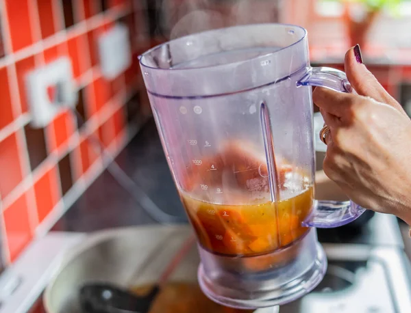 Şef plastik testiyi buğulanmış havuç, zencefil, kırmızı soğan, sarımsak ve stokla dolduruyor. Blender 'da sağlıklı ve besleyici bir ev yapımı çorbaya hazırlanıyor.