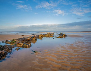 Parlak ama soğuk bir kış gününde, kumlu bir sahilde, uzakta sakin bir deniz olan bir kaya havuzu.