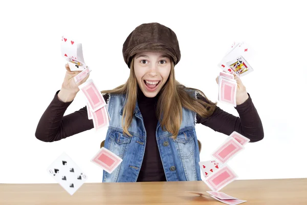 Joyeux jeune fille portant un col roulé brun et gilet en denim avec chapeau sur sa tête jonglant cartes à jouer sur fond blanc et regardant dans la caméra isolé sur blanc — Photo