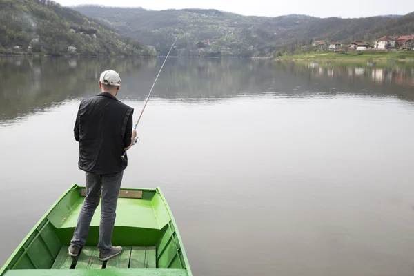 Hombre de pie en un barco verde con la espalda hacia la cámara y la pesca en el lago de la calma — Foto de Stock