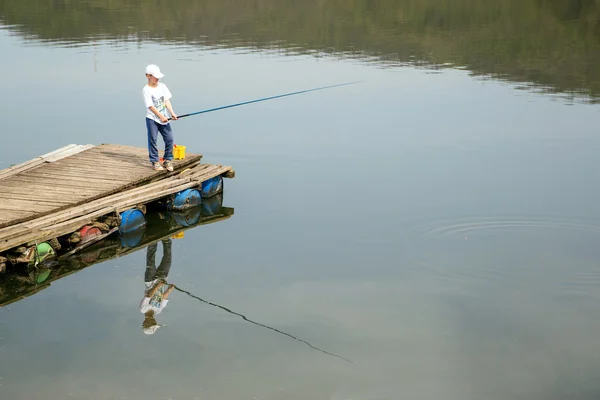 Menino de camisa branca e boné em pé no cais de madeira com uma haste de pesca e pacientemente esperando para pegar um peixe — Fotografia de Stock