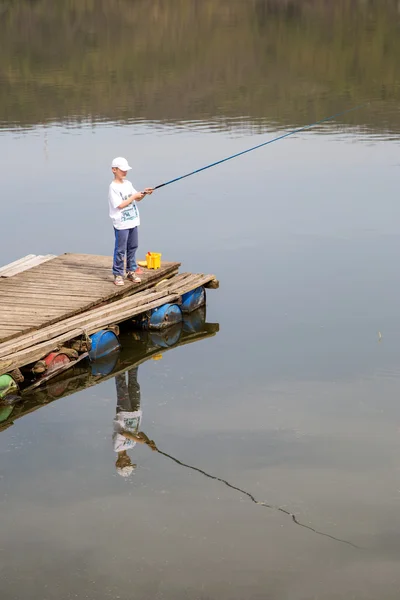 Niño con camisa blanca y gorra de pie en el muelle de madera con una caña de pescar y esperando pacientemente para atrapar un pez — Foto de Stock